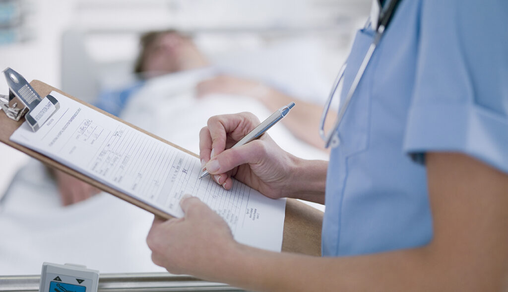 A healthcare professional in scrubs writing on a medical chart near a patient in a hospital bed, indicating clinical evaluation or diagnosis.